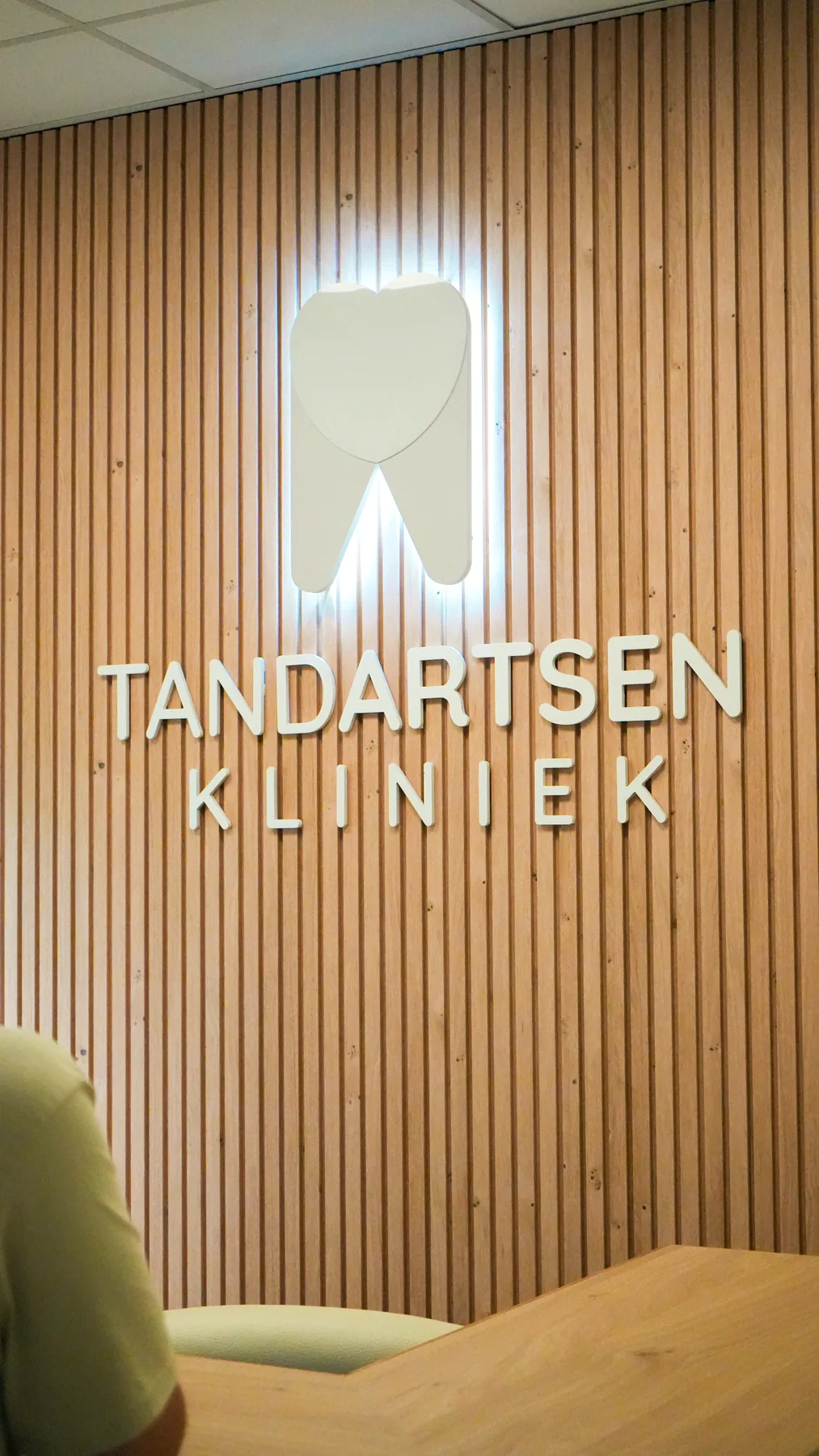 Verlicht logo Tandartsen Kliniek op een houten lamellenwand bij de receptie.