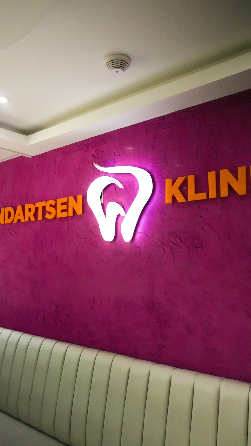 Verlicht logo Tandartsen Kliniek op een paarse wand boven een groene bank.